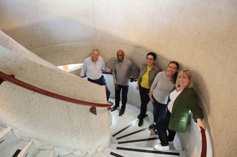 A group of 5 people standing in a modern concrete spiral staircase. All of the people are smiling and looking at the camera